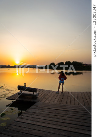 A scenic lakeside view at sunset with a woman standing on a wooden pier, featuring a wooden bench and golden reflections on calm water 125002347