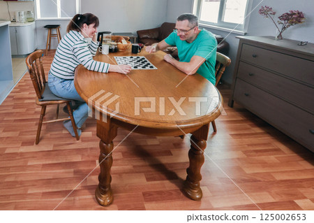 Couple in their 50s is playing checkers at wooden dining table, focused on game, sitting in cozy home with wooden floor and natural light. 125002653