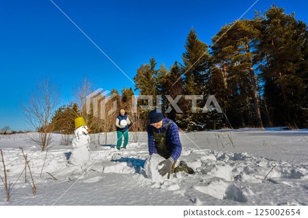 Two people are building a snowman in snowy field on sunny day near forest. 125002654