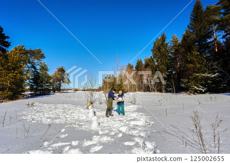 White husband and wife in winter clothes are building snowman, adding yellow bucket on head, possibly for children or holiday fun in snowy landscape. 125002655