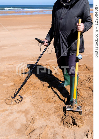 Elderly adult using metal detector on beach wearing black coat and boots 125004087