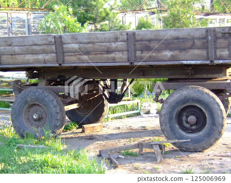 A rustic wooden wagon stands on a gravel surface in a yard, showcasing its robust wheels and structure. Surrounding greenery and buildings create a charming countryside atmosphere. A rustic wooden wagon stands on a gravel surface in a yard, showcasing its robust wheels and structure. Surrounding greenery and buildings create a charming countryside atmosphere. 125006969