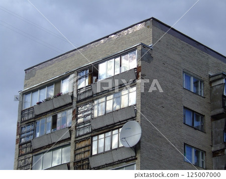 A multi-story building displays significant wear, with broken windows and damaged exterior. Reflections of clouds can be seen on the glass, indicating an overcast day. 125007000