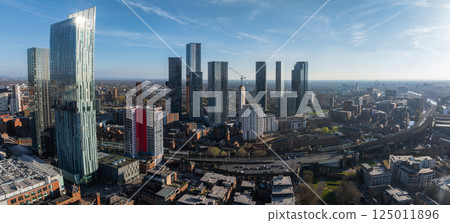 Aerial view of Manchester, UK, featuring Beetham Tower and modern skyscrapers. Roads, railways, and older brick buildings are visible under a clear blue sky. Aerial view of Manchester, UK, featuring Beetham Tower and modern skyscrapers. Roads, railways, and older brick buildings are visible under a clear blue sky. 125011896