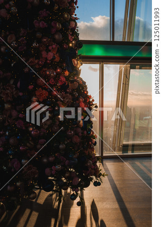 A decorated Christmas tree with ornaments and flowers stands by a window, revealing London's skyline with The Shard at sunset, casting warm shadows. 125011993