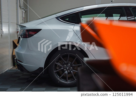 A close up view of a white Tesla Model Y's rear side in a garage with a checkered floor. The scene includes a blurred orange car in the foreground. 125011994
