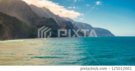 The Napali Coast on Kauai Island features rugged green cliffs descending into turquoise waters, with sunlight highlighting the dramatic landscape. 125012001