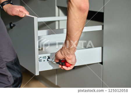 Man assembling kitchen drawer in modern cabinet using screwdriver. Handyman installing and adjusting drawer slide mechanism in contemporary kitchen. Furniture installation Man assembling kitchen drawer in modern cabinet using screwdriver. Handyman installing and adjusting drawer slide mechanism in contemporary kitchen. Furniture installation 125012058