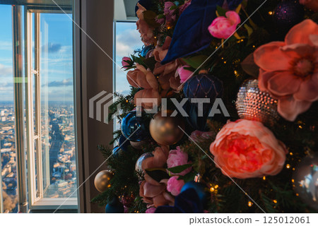 A decorated Christmas tree with large flowers and ornaments stands before a panoramic view of London's cityscape, seen from a high vantage point. A decorated Christmas tree with large flowers and ornaments stands before a panoramic view of London's cityscape, seen from a high vantage point. 125012061