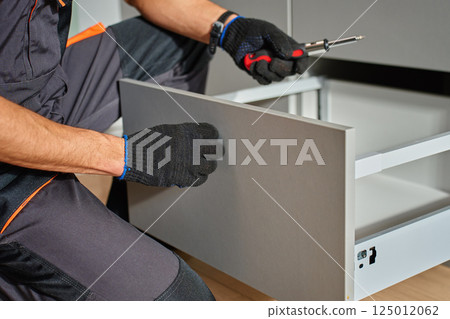 Man assembling kitchen drawer in modern cabinet using screwdriver. Handyman installing and adjusting drawer slide mechanism in contemporary kitchen. Furniture installation Man assembling kitchen drawer in modern cabinet using screwdriver. Handyman installing and adjusting drawer slide mechanism in contemporary kitchen. Furniture installation 125012062