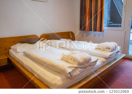 A hotel room featuring a double bed with white linens, brown pillows, and rolled towels. A window with striped curtains reveals a balcony structure. A hotel room featuring a double bed with white linens, brown pillows, and rolled towels. A window with striped curtains reveals a balcony structure. 125012069