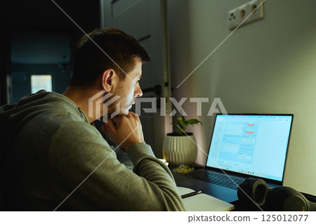 Concentrated freelancer working late at home office. Focused man in hoodie writes notes while working on laptop. Concept of night work 125012077