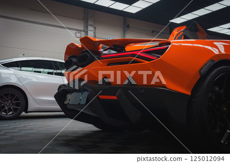 The image features the rear end of an orange Lamborghini Revuelto, highlighting its taillights and dual exhausts. A white car is visible in the background. 125012094