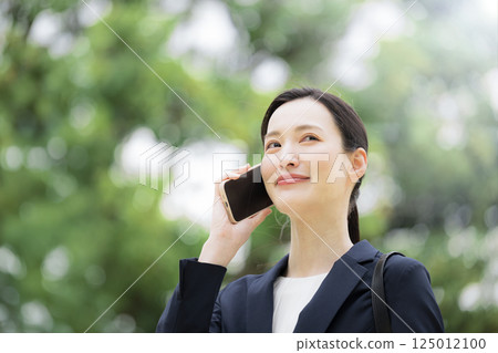 A woman in a business suit talking on her smartphone outdoors in the fresh greenery and looking up. Close-up of an image of sales and outside work A woman in a business suit talking on her smartphone outdoors in the fresh greenery and looking up. Close-up of an image of sales and outside work 125012100