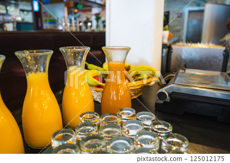 Glass carafes with juices, empty glass cups, fresh bananas, and a stainless steel appliance on a dark countertop in a modern dining area. Glass carafes with juices, empty glass cups, fresh bananas, and a stainless steel appliance on a dark countertop in a modern dining area. 125012175