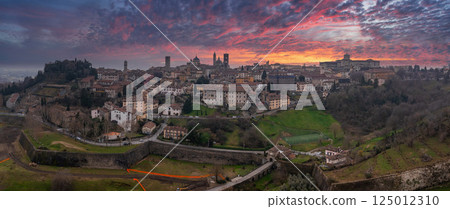 The old town of Bergamo, Italy, features medieval towers, domes, and Venetian walls under a vibrant sunset with pink, orange, and purple hues. The old town of Bergamo, Italy, features medieval towers, domes, and Venetian walls under a vibrant sunset with pink, orange, and purple hues. 125012310