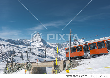 A bright orange Gornergrat Railway train crosses a snow covered stone bridge in Zermatt, Switzerland, with the Matterhorn and alpine scenery in view. 125012322