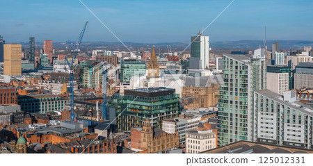 Aerial view of Manchester, UK, featuring the Town Hall clock tower, skyscrapers, historic and modern buildings, cranes, and distant rolling hills. 125012331