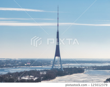 The Riga TV Tower stands on an island in the Daugava River, surrounded by snow and frozen water, with Riga's cityscape and bridges in the background. 125012379