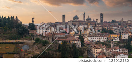View of Bergamo's old town in Italy during sunset, featuring the Campanone, Basilica of Santa Maria Maggiore, medieval architecture, and lush greenery. View of Bergamo's old town in Italy during sunset, featuring the Campanone, Basilica of Santa Maria Maggiore, medieval architecture, and lush greenery. 125012380