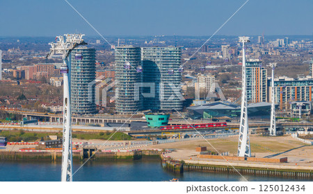 Aerial view of London's Emirates Air Line cable cars, tall white towers, modern high rise buildings, a red train, and waterfront construction activity. Aerial view of London's Emirates Air Line cable cars, tall white towers, modern high rise buildings, a red train, and waterfront construction activity. 125012434