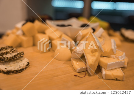 An assortment of cheeses on a wooden platter, featuring brie wedges, herb coated slices, and cubes. A bowl of dark olives and blurred fruits are in the background. 125012437