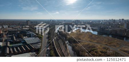 Aerial view of Manchester, UK, showing a river, modern skyscrapers, industrial buildings, railway, road, open land, and construction cranes under a clear sky. 125012471