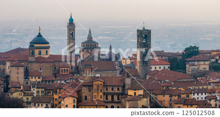 View of Bergamo's old town featuring the Basilica of Santa Maria Maggiore, Campanone, and Cappella Colleoni amid terracotta roofs and a hazy sunset. 125012508