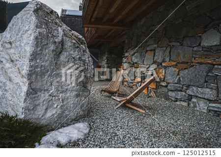 Two wooden lounge chairs on gravel near a large boulder, with a stone and wood building in Alpine style and snow on the ground in a serene setting. Two wooden lounge chairs on gravel near a large boulder, with a stone and wood building in Alpine style and snow on the ground in a serene setting. 125012515