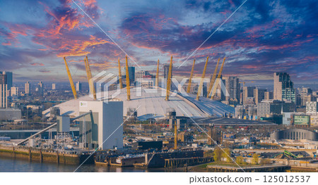 Aerial view of the O2 Arena with its white dome and yellow masts, surrounded by modern buildings, the Thames River, and urban development in London. 125012537