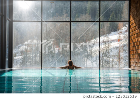 A serene indoor pool with clear water, a person relaxing by the edge, and a floor to ceiling window revealing snowy slopes and alpine trees. 125012539