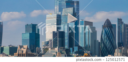 The image captures London's skyline, highlighting the Gherkin and Leadenhall Building. Modern skyscrapers with glass facades stand under a clear blue sky. 125012571