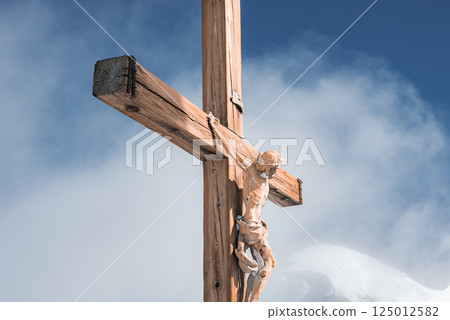 A detailed wooden crucifix of Jesus Christ stands against a clear blue sky, with snow covered mountain peaks and soft clouds in the Zermatt Alps. 125012582