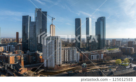 Aerial view of Manchester, UK, featuring modern skyscrapers, a construction crane, the River Irwell, and historic brick buildings under a clear blue sky. 125012583