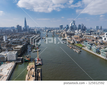 Aerial view of London with the River Thames, Tower Bridge, The Shard, and modern skyscrapers under a partly cloudy sky with boats on the river. 125012592