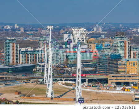 Aerial view of London featuring Emirates Air Line cable cars over the Thames, modern buildings, the O2 Arena, and urban construction areas. 125012611