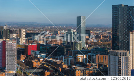 Aerial view of Manchester, UK, showcasing modern skyscrapers, mixed architecture, and rolling hills in the background on a sunny day. 125012612