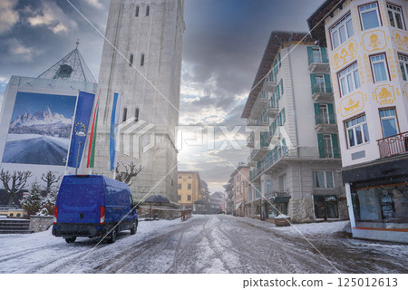 A snowy street in Cortina d'Ampezzo, Italy, featuring a tall bell tower, a mural of mountains, charming alpine buildings, and a parked blue van. A snowy street in Cortina d'Ampezzo, Italy, featuring a tall bell tower, a mural of mountains, charming alpine buildings, and a parked blue van. 125012613