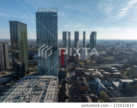 Aerial view of Manchester, UK, showcasing skyscrapers like Beetham Tower, modern and historic architecture, streets, and a unique roof structure. 125012614