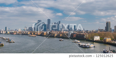 Aerial perspective of London's Canary Wharf skyline with modern skyscrapers, the River Thames with boats, and a mix of historic and modern buildings. 125012625