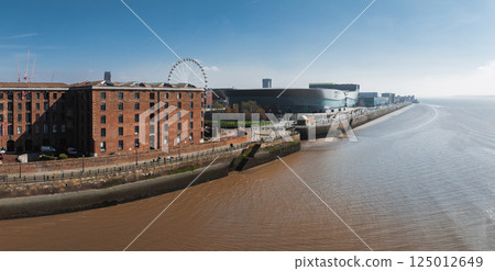 Aerial view of Liverpool showcasing Albert Dock's red brick warehouses, the Liverpool Wheel, MandS Bank Arena, and the River Mersey under a clear sky. 125012649