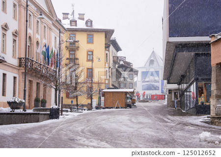 A snowy street in Cortina d'Ampezzo, Italy, featuring colorful buildings, flags, bare trees, and a glass roofed structure with a mountain mural. A snowy street in Cortina d'Ampezzo, Italy, featuring colorful buildings, flags, bare trees, and a glass roofed structure with a mountain mural. 125012652