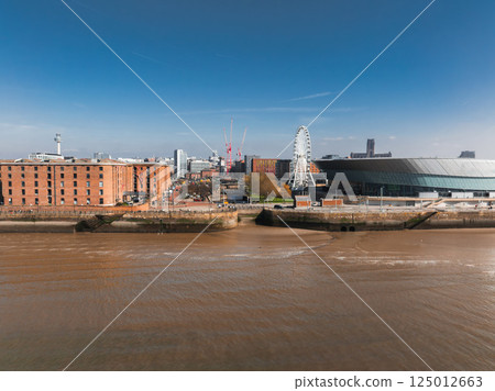Aerial view of Liverpool with Albert Dock's red brick warehouses, MandS Bank Arena, the Liverpool Wheel, and the River Mersey under a clear blue sky. Aerial view of Liverpool with Albert Dock's red brick warehouses, MandS Bank Arena, the Liverpool Wheel, and the River Mersey under a clear blue sky. 125012663