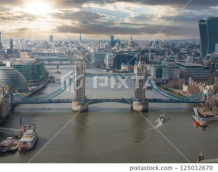 Tower Bridge spans the River Thames with City Hall, the Walkie Talkie, St. Paul's Cathedral, and the Shard visible under a partly cloudy sky. 125012670