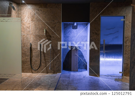 Spa area featuring stone walls, modern showers, a blue lit stone basin, and a glass door leading to a steam sauna in a high end Italian Alps hotel. 125012791