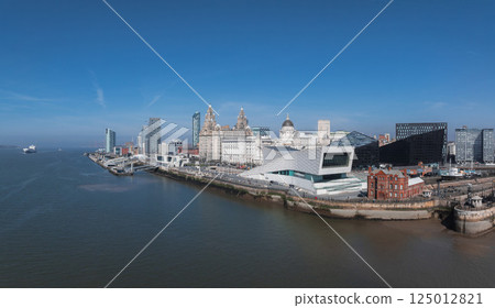 Aerial view of Liverpool's waterfront featuring the Three Graces, the Museum of Liverpool, and the River Mersey under a clear blue sky. 125012821