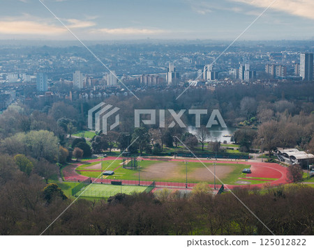 Aerial view of a London park featuring a red running track, grassy field, trees, and a small lake, with urban buildings visible in the distance. 125012822