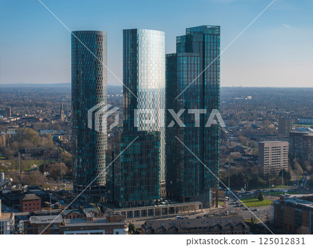 Aerial view of Manchester, UK, featuring three reflective glass skyscrapers surrounded by smaller buildings, greenery, and a clear blue sky. Aerial view of Manchester, UK, featuring three reflective glass skyscrapers surrounded by smaller buildings, greenery, and a clear blue sky. 125012831
