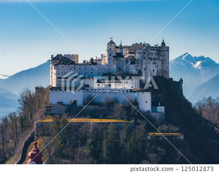 Hohensalzburg Fortress atop Festungsberg hill in Salzburg, Austria, with white walls, towers, forested slopes, and snow capped mountains in the background. 125012873
