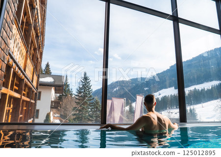 A person relaxes in an indoor pool with large windows, showcasing the Italian Alps, a wooden chalet, evergreen trees, and snowy hills in the background. A person relaxes in an indoor pool with large windows, showcasing the Italian Alps, a wooden chalet, evergreen trees, and snowy hills in the background. 125012895